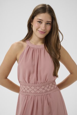 Fiatal nő hosszú barna hajjal, ujjatlan poros rózsaszín ruhát visel csipkés derékpánttal, mosolyogva a kezeit a háta mögött tartva.