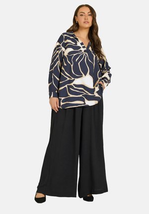 Mujer que lleva una blusa holgada con estampado azul marino y beige, pantalones negros de pierna ancha y zapatos negros, de pie con una mano en el bolsillo.