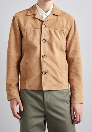 Personne portant une veste en daim marron boutonnée par-dessus une chemise blanche avec un pantalon vert visible des hanches jusqu’au cou.