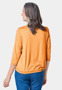 Orange 3/4-Ärmel-Oberteil mit gerafftem Saum, glatter Textur und lockerer Passform, kombiniert mit blauen Jeans.