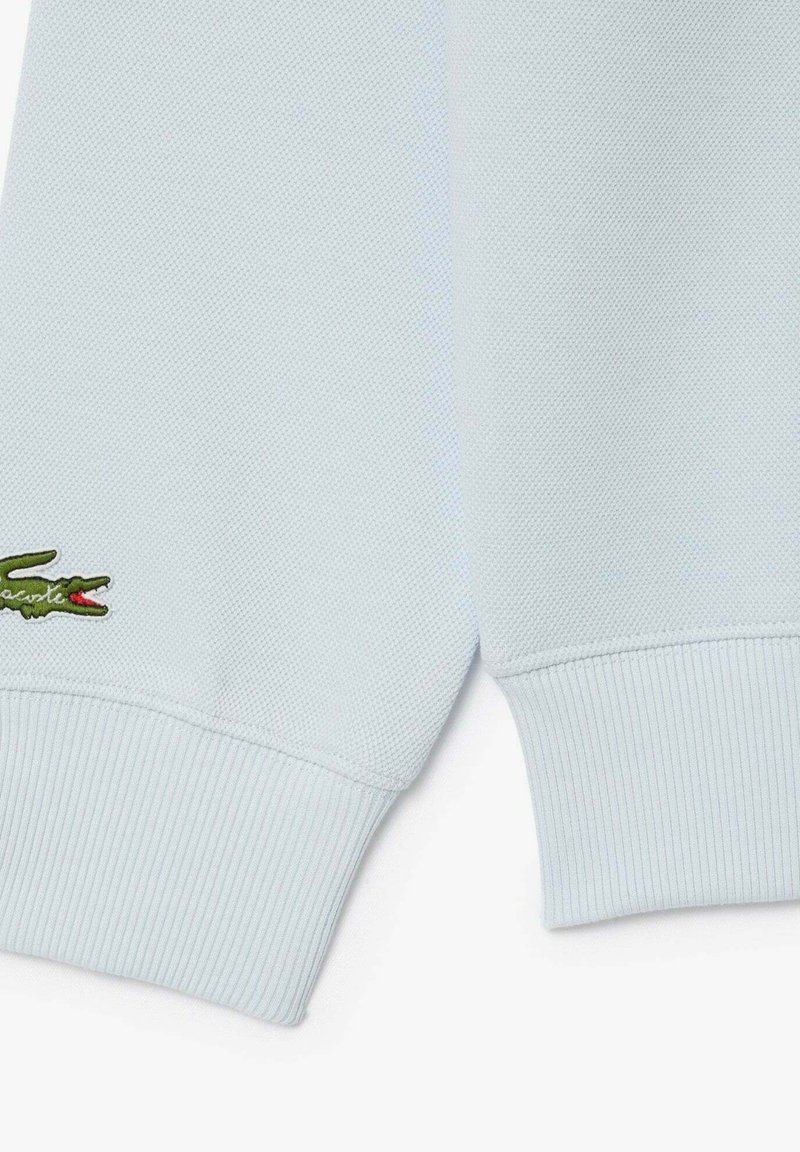 Tissu bleu clair texturé avec des poignets côtelés ; présente un logo de crocodile brodé vert et rouge.