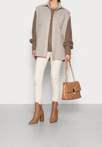 Beżowa koszula-kurtka z mieszanki wełny z dwoma kieszeniami, brązowy sweter dzianinowy, białe skinny jeansy i beżowe botki na obcasie. Duża beżowa torebka z łańcuszkowym paskiem.