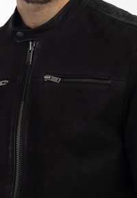 Schwarze Lederjacke mit vertikalem Frontreißverschluss, hohem Kragen mit Druckknopf und zwei horizontalen, mit Reißverschluss versehenen Brusttaschen.