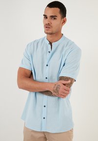 Camisa de botones de manga corta de color azul claro, hecha de un tejido texturizado, con cuello mao y botones oscuros en el frente.