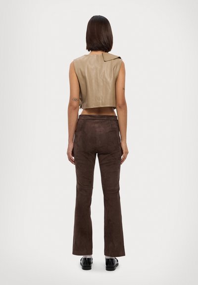 Beige Lederoberteil mit einem einzelnen Schulterlapel, kombiniert mit braunen Wildlederhosen im Schlag. Schwarze Schuhe runden das Outfit ab.