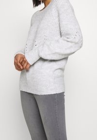 Pull en maille gris clair avec un ourlet et des poignets côtelés. Présente une coupe décontractée et un design en tricot ouvert avec de petits détails en œillets.