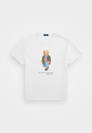 Balta kokvilnas T-kreklu ar karikatūru lāci ikdienas drēbēs, ieskaitot zaļu džemperi, svītrainu krekliņu un džinsus, ar zīmola nosaukumu zem tā.