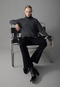 Grijze coltrui gecombineerd met zwarte flared broek. Model zit op een metalen stoel. Zwarte hoge hakken voltooien de outfit.