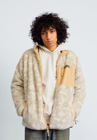 Fleecejacke in Cremefarbe mit einem sandfarbenen vorderen Paneel, Reißverschlusstasche und strukturierter Oberfläche; verfügt über eine Kapuze und Fronttaschen.