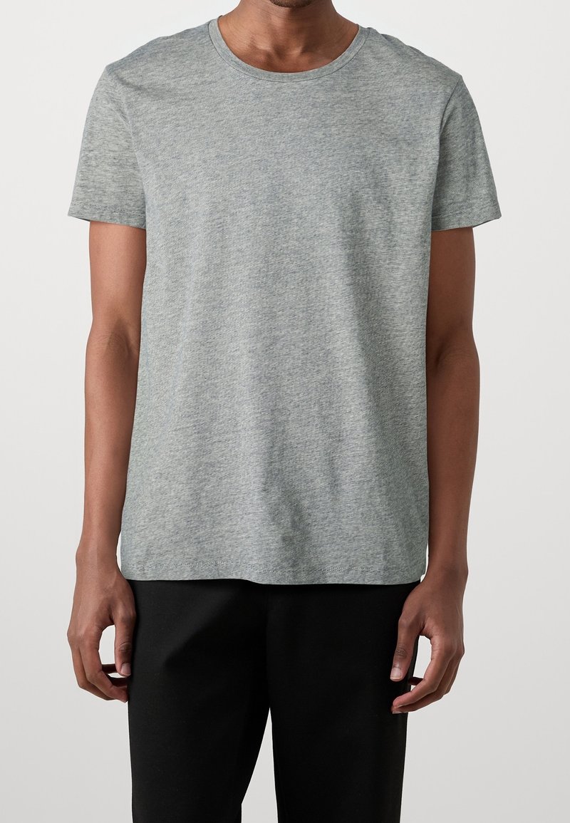 Homme portant un t-shirt gris uni à manches courtes et un pantalon noir, debout devant un fond neutre, les bras détendus le long du corps.