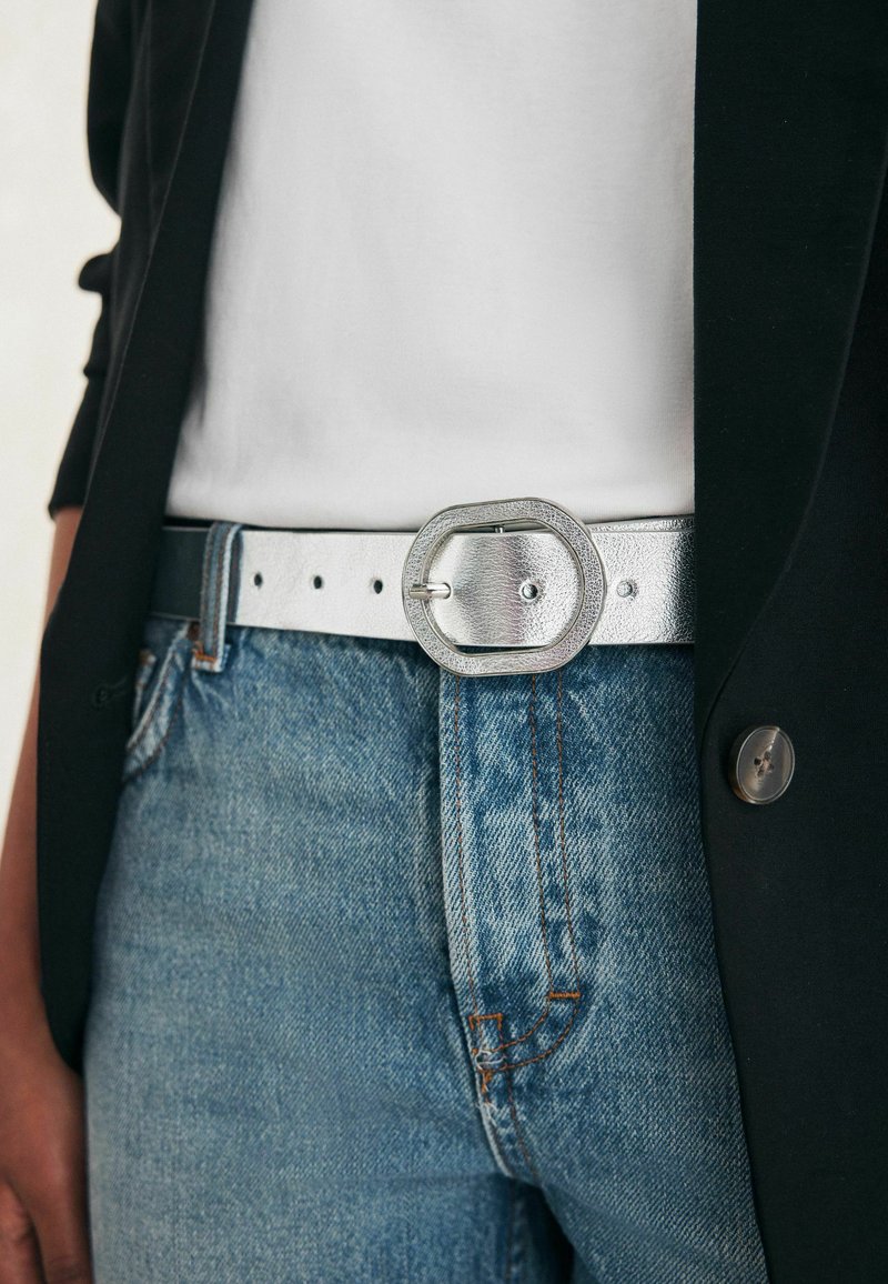 Sølv læderbælte med en ottekantet spænde, båret over denim jeans og en hvid skjorte under en sort jakke. Struktureret finish, flere huller.