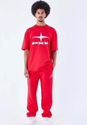 Homme aux cheveux afro portant un t-shirt rouge avec le logo PXPF et un pantalon assorti, debout avec une main dans la poche et des baskets blanches.