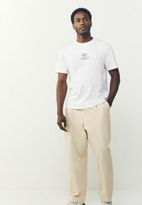 Hvid bomulds T-shirt med et lille rødt grafisk design på brystet, parret med beige plisserede bukser og hvide sneakers.