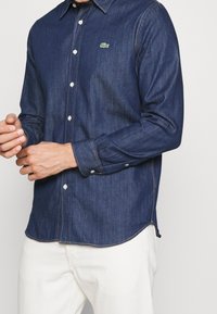 Man som bär en mörkblå denimskjorta med knappar och långa ärmar, samt vita byxor, och justerar manschetten med händerna synliga.