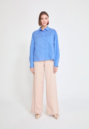 Camisa azul de botones con rayas verticales, ajuste holgado y cuello, combinada con pantalones anchos de color claro y tacones con estampado.