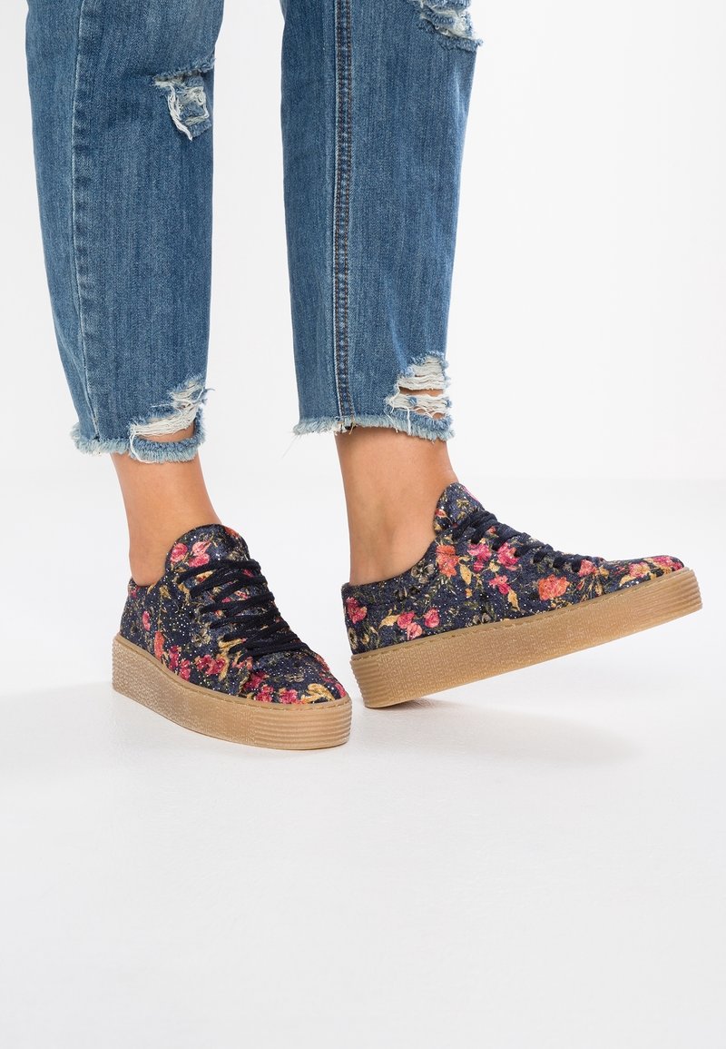 Baskets à motifs floraux sur fond sombre, avec des lacets noirs et une semelle en caoutchouc texturée de couleur tan. Portées avec un jean bleu déchiré.