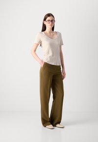 Tricou gri deschis cu decolteu în V, croială lejeră; pantaloni oliv verzi, design cu picioare largi; încălțăminte minimalistă; modelul stă cu mâinile în buzunare.