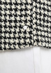 Tela con patrón de pata de gallo en blanco y negro con un acabado texturizado, que presenta una pequeña etiqueta metálica de la marca en el borde. Fondo en un suave blanco.