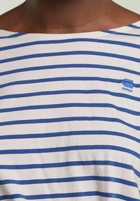 Camiseta a rayas con líneas horizontales azules y blancas, hecha de una tela suave, con un pequeño logotipo azul en el lado izquierdo.