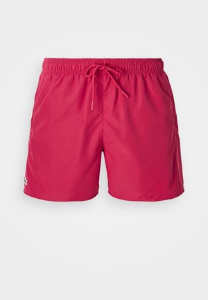 Shorts de bain rouges avec une taille élastique et un cordon de serrage. Présente un petit logo vert sur le bas à gauche. Matériau lisse et léger.