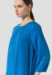 Blaue Bluse mit rundem Ausschnitt, langen Puffärmeln und einem schlichten Muster. Der Stoff wirkt glatt und leicht mit einer lockeren Passform.