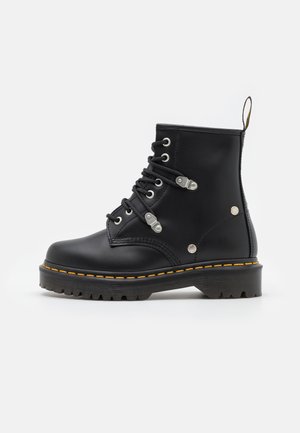 Bottines noires en cuir avec un bout rond, œillets en argent et lacets noirs. Présentent une semelle texturée avec des coutures jaunes sur le bord.