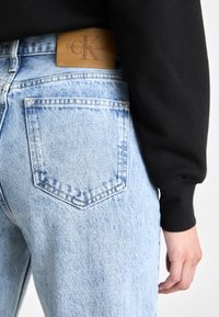 Unos vaqueros de denim azul claro presentan un corte relajado, con una etiqueta de cuero marrón de Calvin Klein en la cintura y un diseño sencillo en el bolsillo trasero.