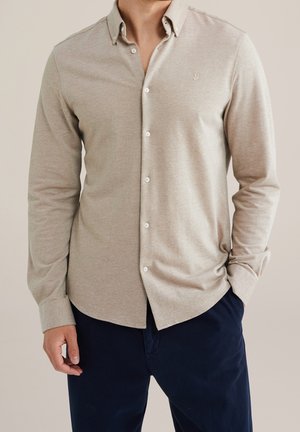 Mężczyzna ubrany w beżową koszulę na długi rękaw zapinaną na guziki z małym wyszywanym logo na piersi oraz ciemnogranatowe spodnie.