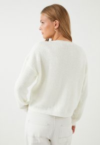 Weißer gerippter Pullover mit lockerer Passform und überschnittenen Schultern, ausgestattet mit langen Ärmeln und einem weiten Ausschnitt, kombiniert mit hellen Hosen.