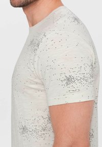 Camiseta de manga corta color beige claro con un patrón de texto negro disperso. Textura suave con una etiqueta en la manga.