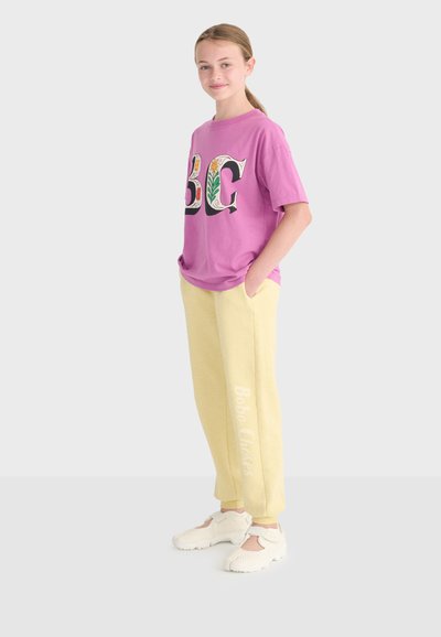 Jeune fille debout les mains dans les poches, portant un t-shirt graphique oversize violet à motifs floraux, un pantalon de jogging jaune et des baskets blanches.