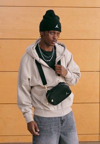 Sweatshirt bege com fecho éclair, t-shirt preta, jeans cinzentos e uma pequena bolsa tiracolo preta. Gorro preto com um logótipo. Colar de corrente prateada.