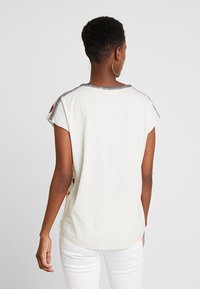 Kortærmet elfenbensfarvet t-shirt med metallisk grå kant langs kraven og skuldrene. Blød tekstur og rund kant med mønstrede detaljer.