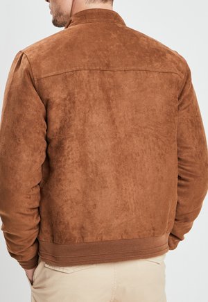 Mann trägt eine braune Wildlederjacke mit gerippten Bündchen und Bund, steht mit den Händen in den Taschen, beige Hose ist von hinten sichtbar.