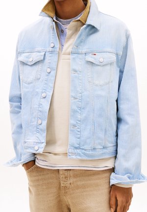 Mann trägt eine hellblaue Jeansjacke mit Nadelstreifen über einem beigen Hemd und hellbraunen Hosen, mit der linken Hand in der Tasche und rechtem Arm entspannt.