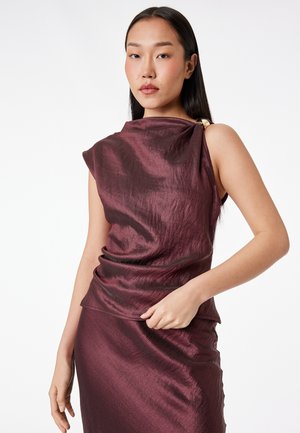 Kobieta o długich czarnych włosach, ubrana w błyszczącą, ciemnofioletową bluzkę bez rękawów i pasującą spódnicę na jednolitym białym tle.