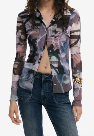 Kvinde iført en langærmet, abstrakt mønstret skjorte med knapper, delvist åben, kombineret med mørkeblå jeans og et guldb armbånd på højre håndled.