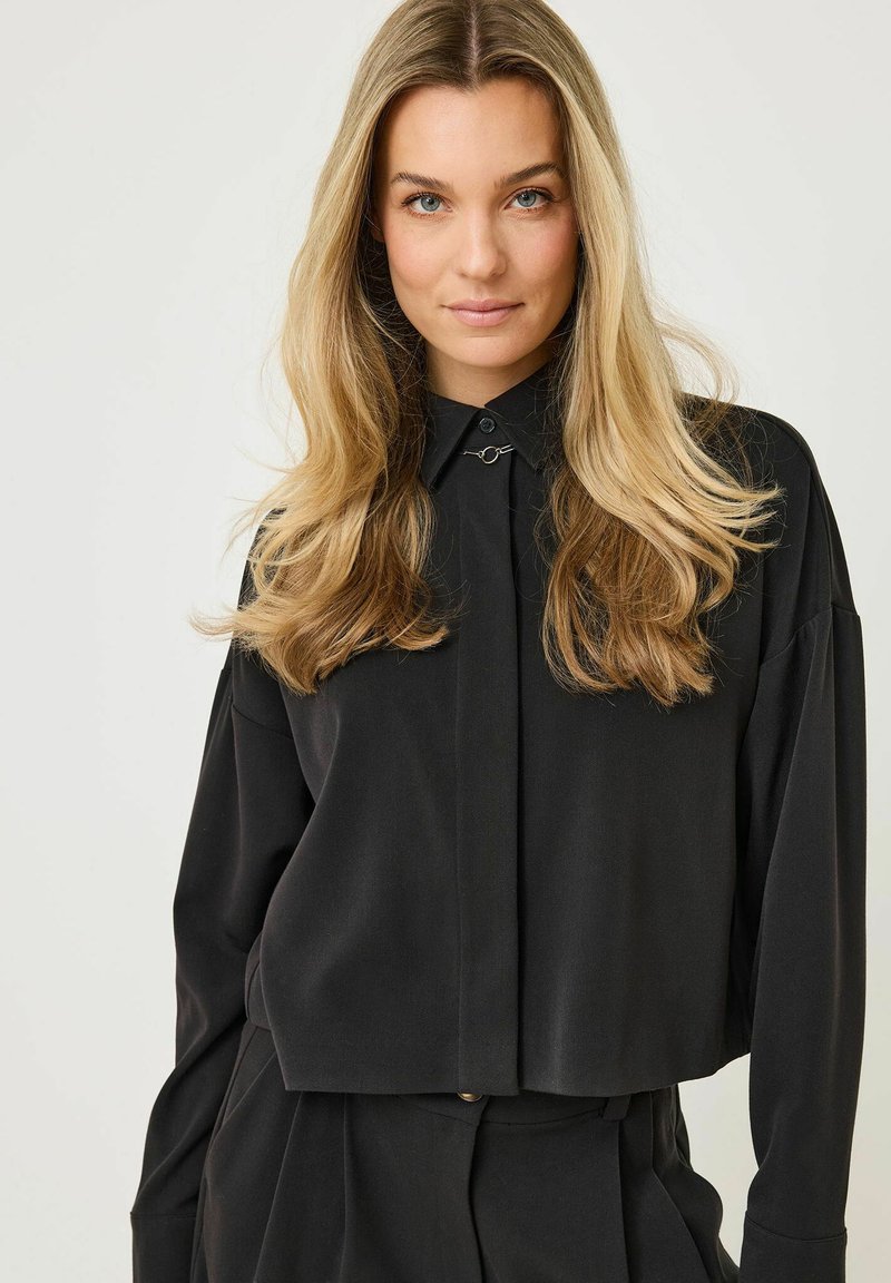Chemise noire courte à manches longues, fabriquée en tissu lisse. Présente une fermeture à boutons sur le devant et une pince argentée décorative au col.
