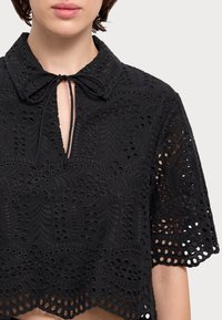 Melns izšūts tops ar apkakli, atslēgas caurumu detaļu un īsām piedurknēm. Iezīmējas sarežģīti izgriezti raksti un viļņota apakšmala.