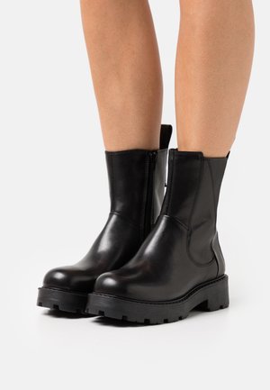 Bottes noires en cuir à la cheville avec des semelles épaisses, portées sur des jambes nues, devant un fond blanc uni.