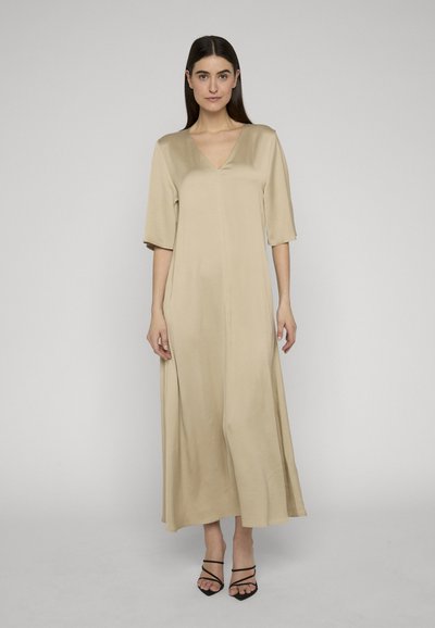 Femme aux cheveux noirs et raides portant une robe midi beige à col en V, ample, avec des manches trois-quarts, et des talons noirs à lanières, debout sur un sol gris.