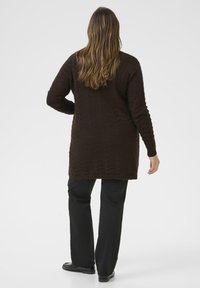 Brauner gestrickter langer Cardigan mit einem strukturierten Zickzackmuster, kombiniert mit schwarzen Hosen. Das Outfit hat lange Ärmel und eine lockere Passform.