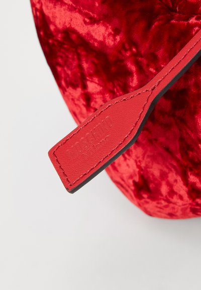 Sac en velours rouge avec une texture brillante, présentant une étiquette en cuir noir et rouge en relief avec le mot "Moschino." Forme ronde, surface douce.