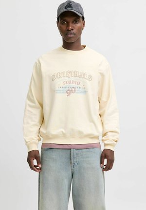 Creme sweatshirt med tekstdesign i falmet rød og blå, rund hals, ribbede manchetter, lagd over en pink skjorte, parret med lyse jeans.