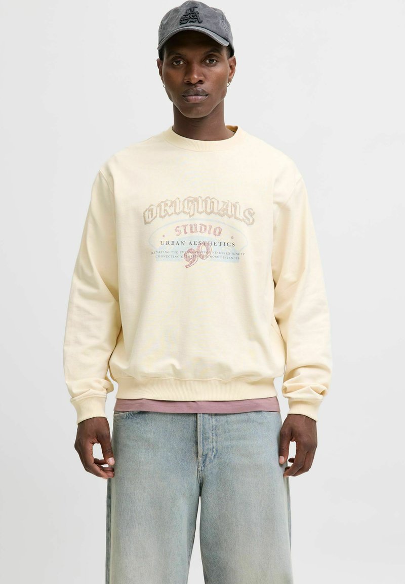 Creme sweatshirt med tekstdesign i falmet rød og blå, rund hals, ribbede manchetter, lagd over en pink skjorte, parret med lyse jeans.
