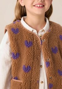 Gilet en polaire marron avec des motifs de cœurs violets, doté d'une fermeture à pression et d'un bord beige. Porté par-dessus une chemise blanche à col avec des boutons.