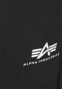 Svart tyg med vertikal ribbad struktur och en vit logotyp med stiliserade bokstäver "A" och "I", vilket indikerar varumärket Alpha Industries.