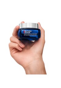 Niebieski szklany słoik z metalową pokrywką, oznaczony etykietą "Biotherm Homme Force Supreme Face Reshaper". Trzymany w dłoni o gładkiej teksturze skóry.