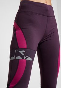 Leggings sportivi di un viola scuro con dettagli rosa e logo grigio. Presentano un tessuto liscio e elastico e un alto punto vita per il massimo comfort.