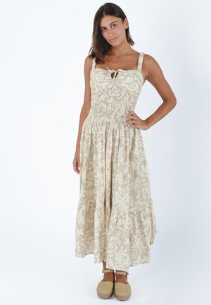 Giovane donna in piedi con mano sul fianco, indossa un abito lungo senza maniche con motivo floreale beige e bianco e espadrillas beige.